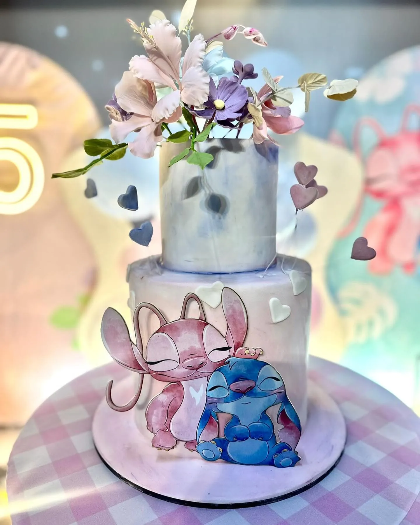 Bolo de aniversário de dois andares com tema Stitch e Angel em tons pastéis, com corações e bonecos se abraçando na base.