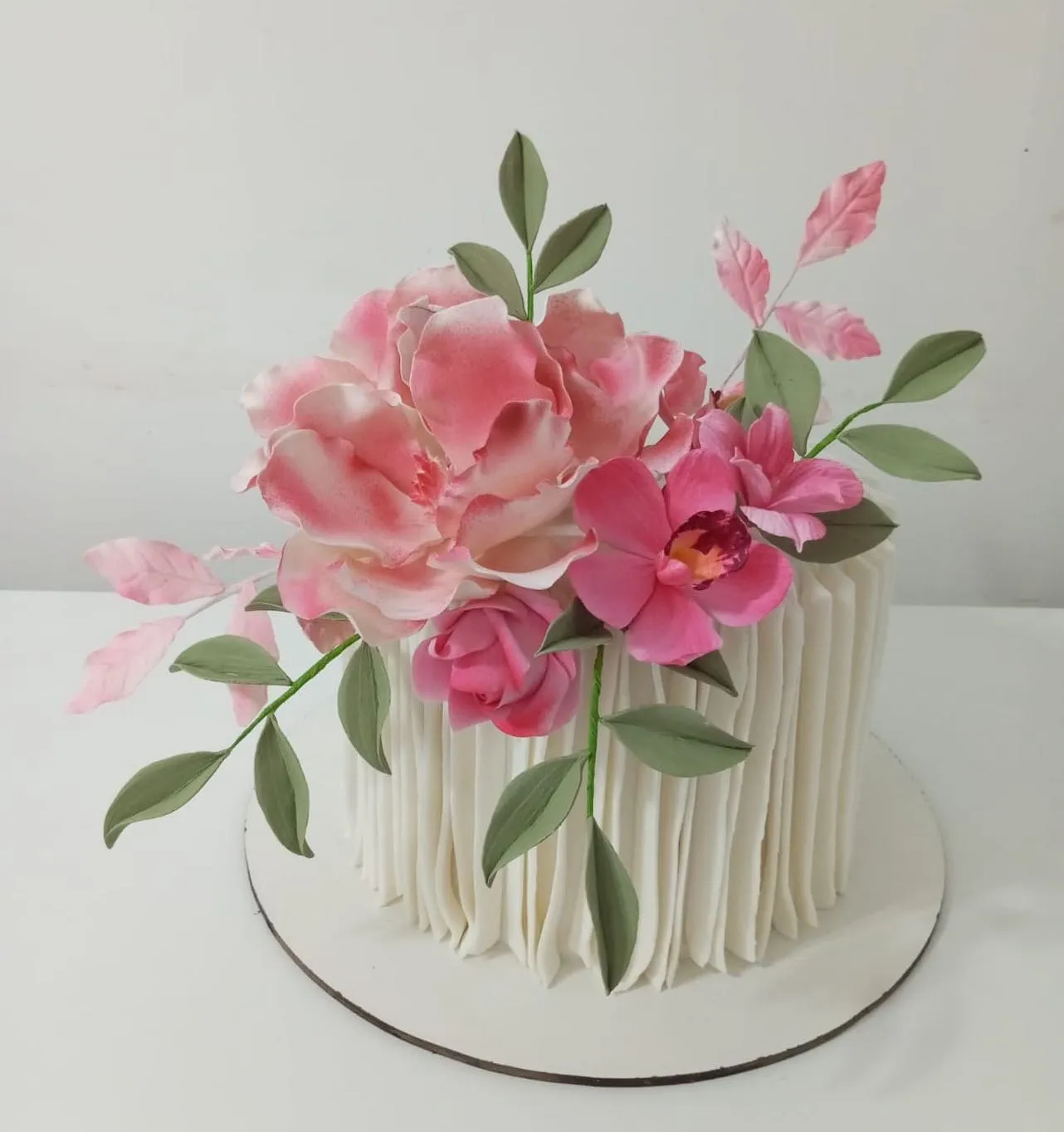 Bolo de confeitaria alto e branco com textura plissada e um buquê de flores comestíveis em tons de rosa e pink no topo.