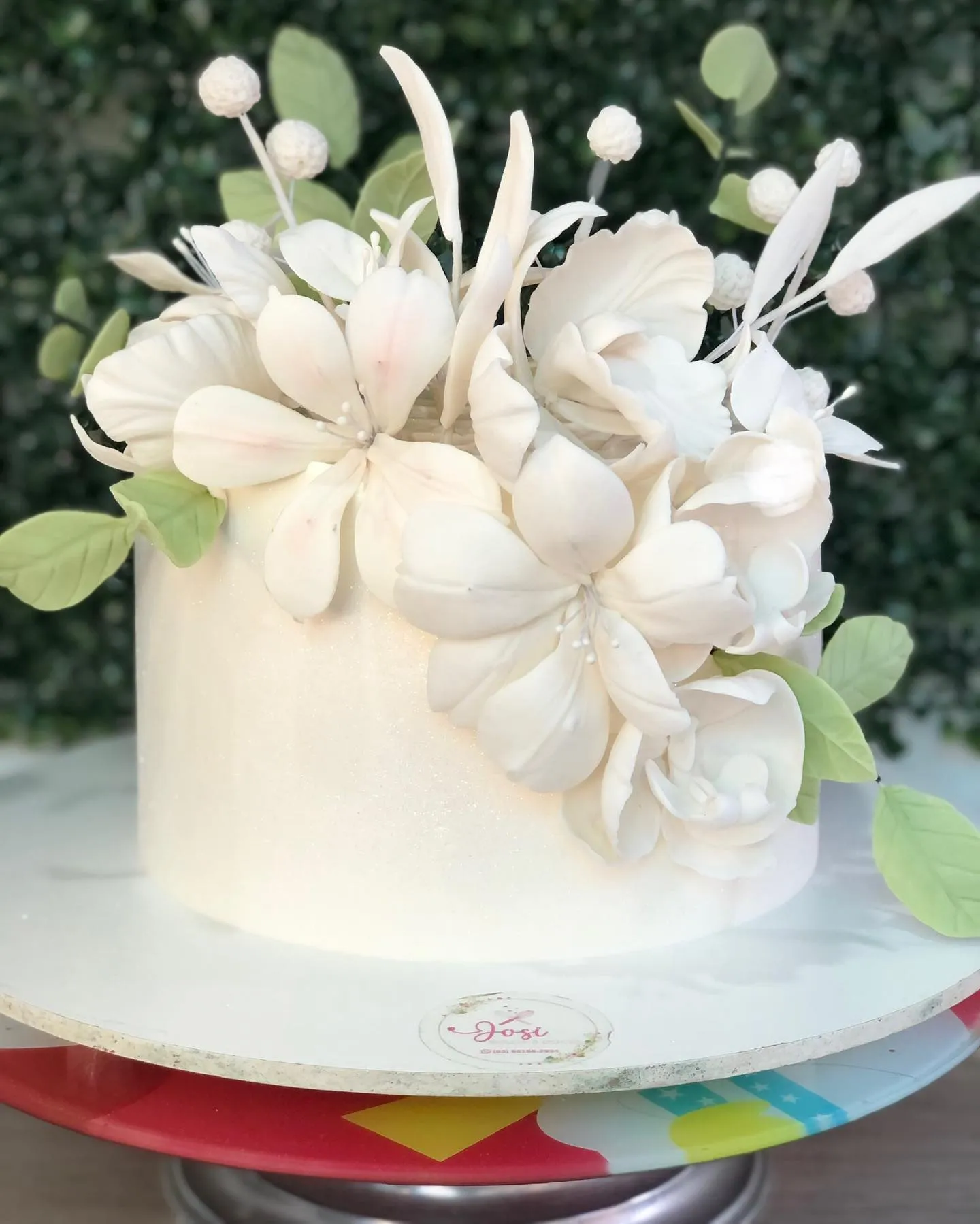 Bolo de aniversário branco e cilíndrico com cobertura brilhante, decorado no topo com um buquê de flores de açúcar