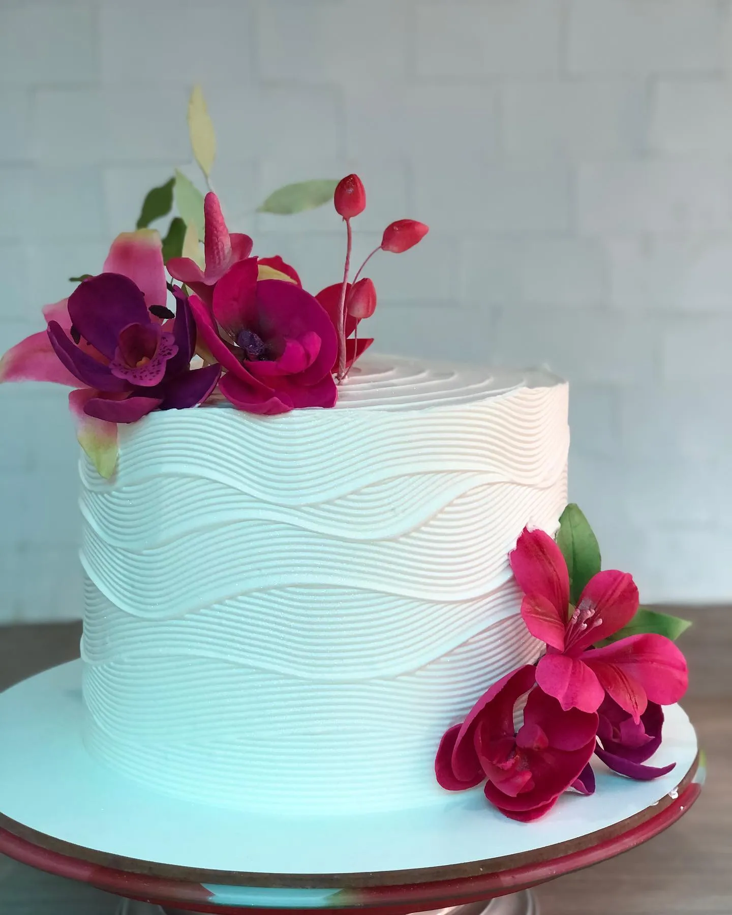 Bolo branco de um andar com textura ondulada sobre um suporte vermelho, adornado com orquídeas roxas e flores magenta.