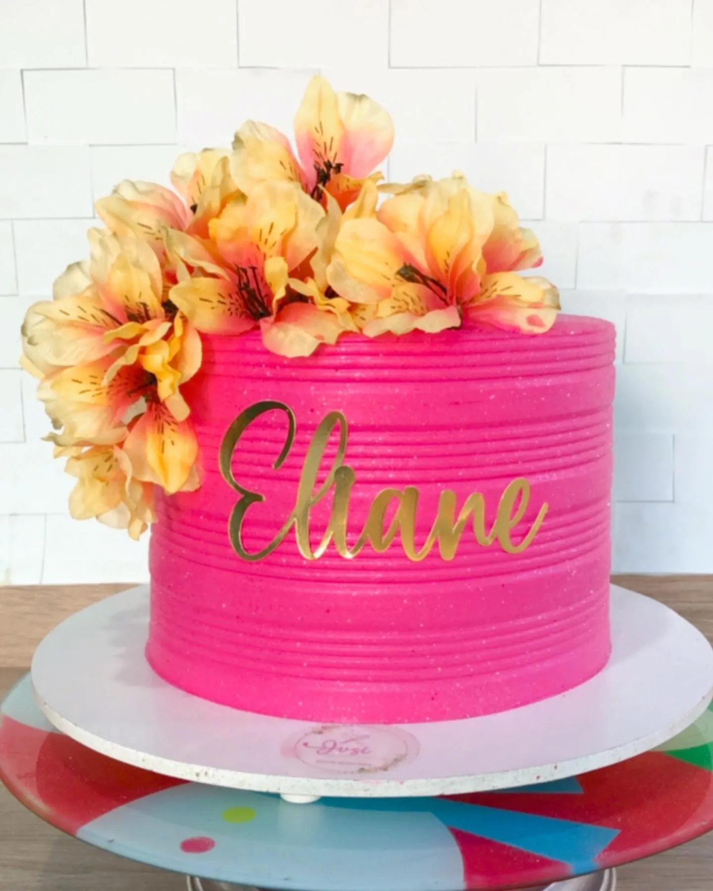Bolo de aniversário alto e redondo, com cobertura rosa-choque, decorado com flores amarelas e o nome Eliane em dourado.