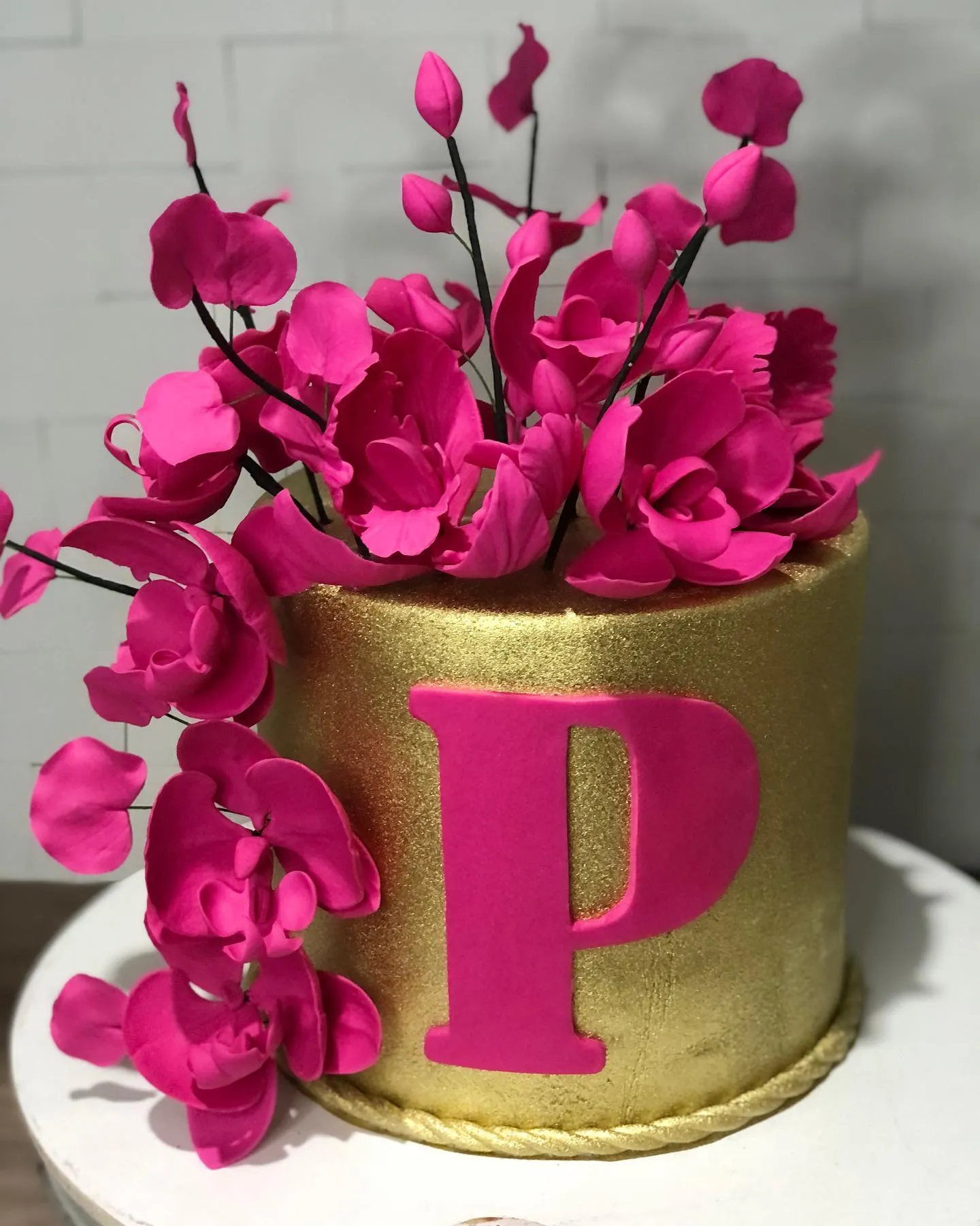 Bolo de festa cilíndrico dourado com a letra 'P' em rosa pink na frente, decorado com um arranjo de orquídeas no topo.