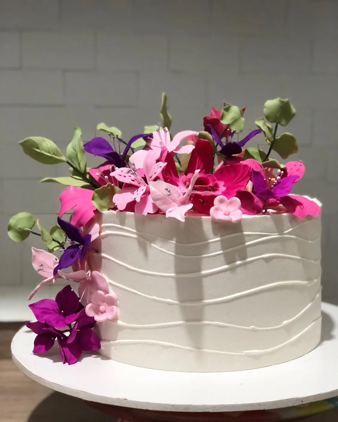 Um bolo branco cilíndrico com textura ondulada, decorado com flores rosa, fúcsia e roxas em cascata
