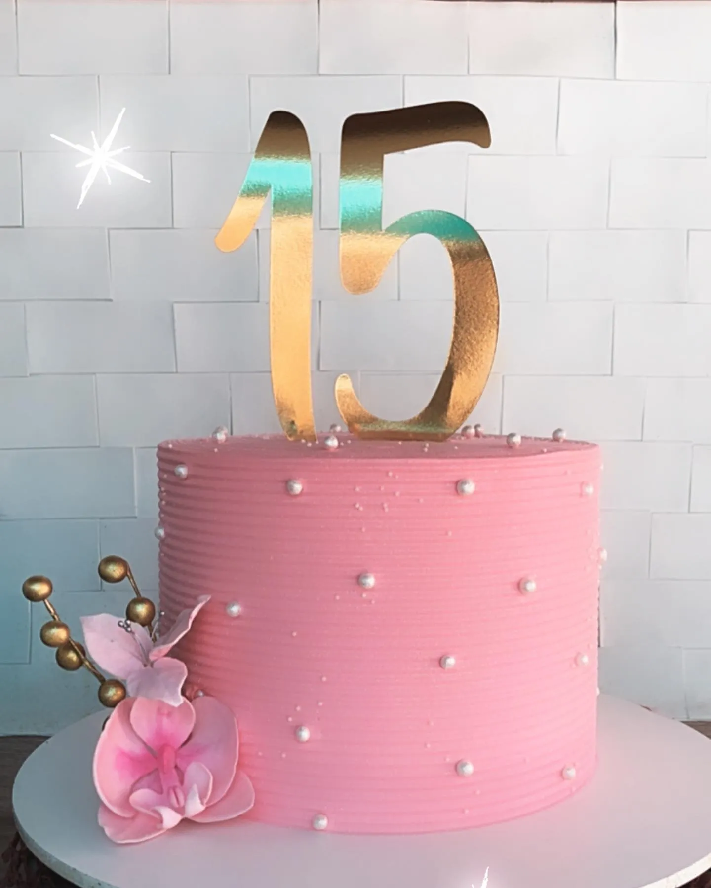 Bolo de festa feminino, alto e redondo, na cor rosa, com pérolas brancas, um topo dourado e um arranjo floral na base.