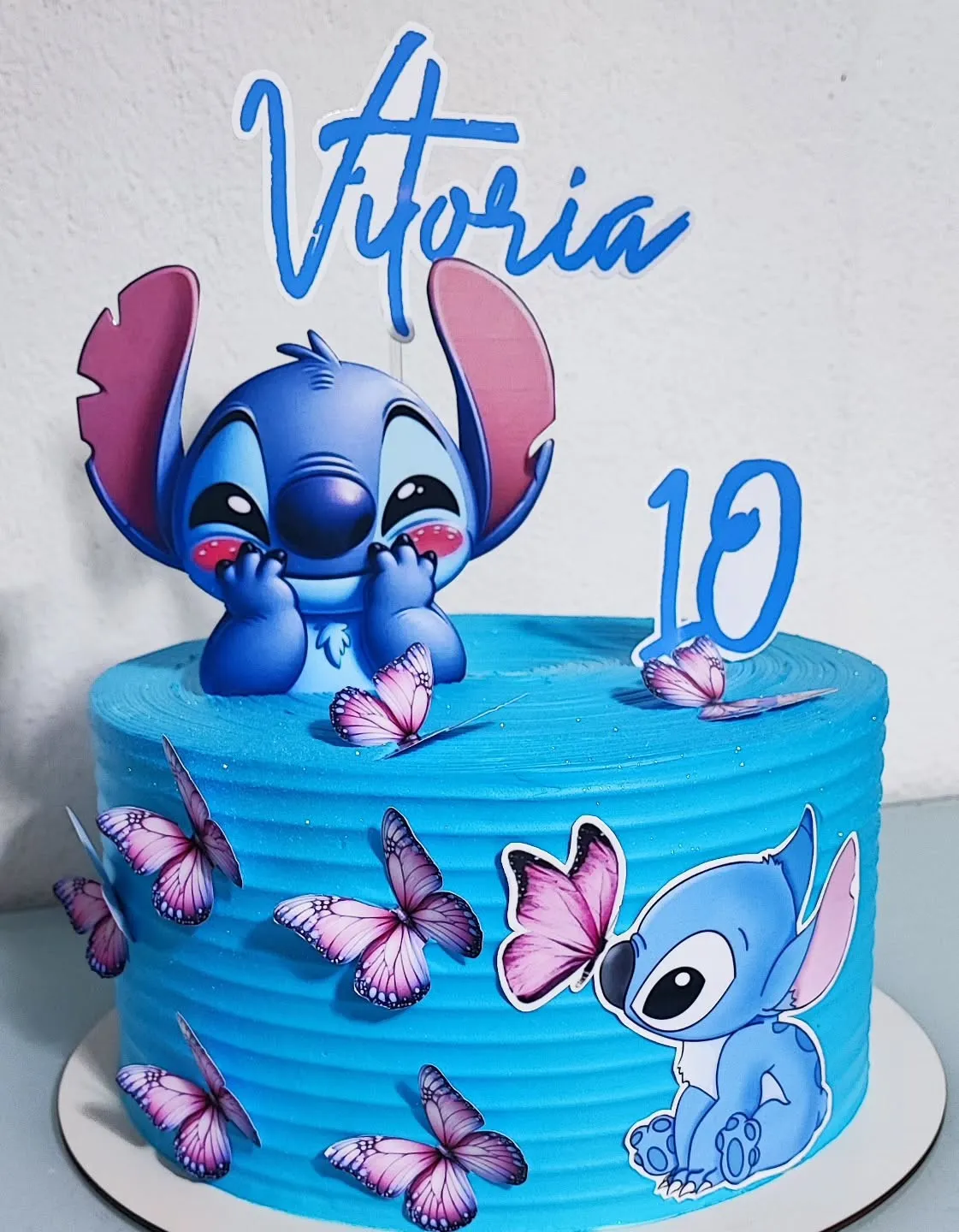 Um bolo de festa redondo com cobertura azul e o tema do Stitch. A decoração mostra o Stitch cercado de borboletas.