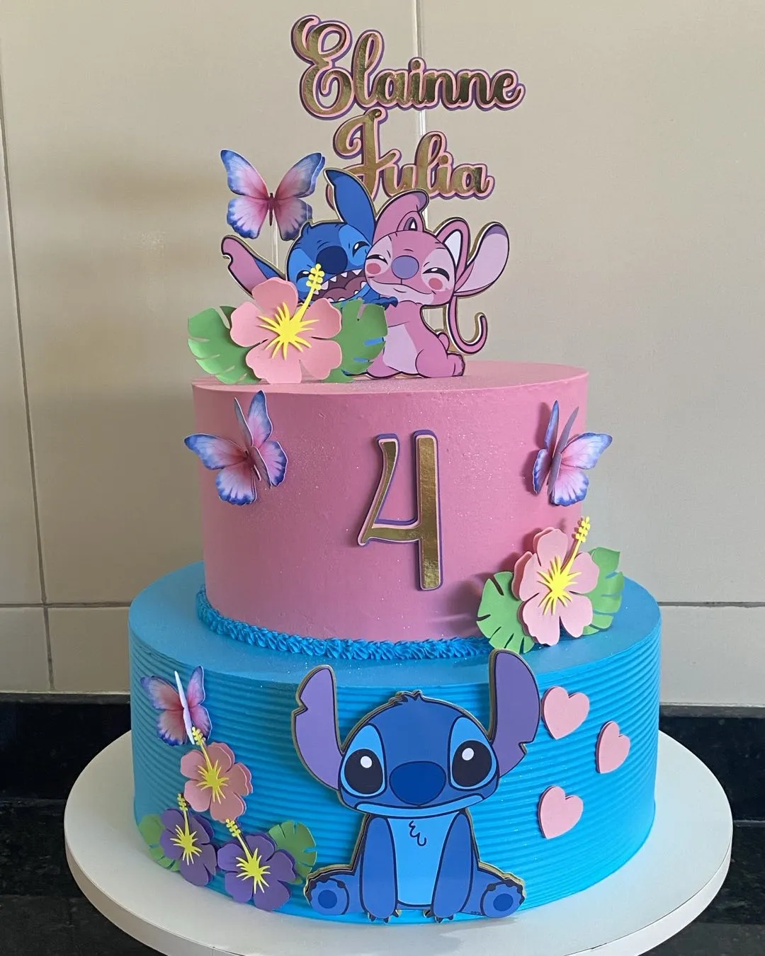 Bolo de festa de dois andares com tema de Stitch e Angel, apresentando um topo com casal de bonecos e flores.