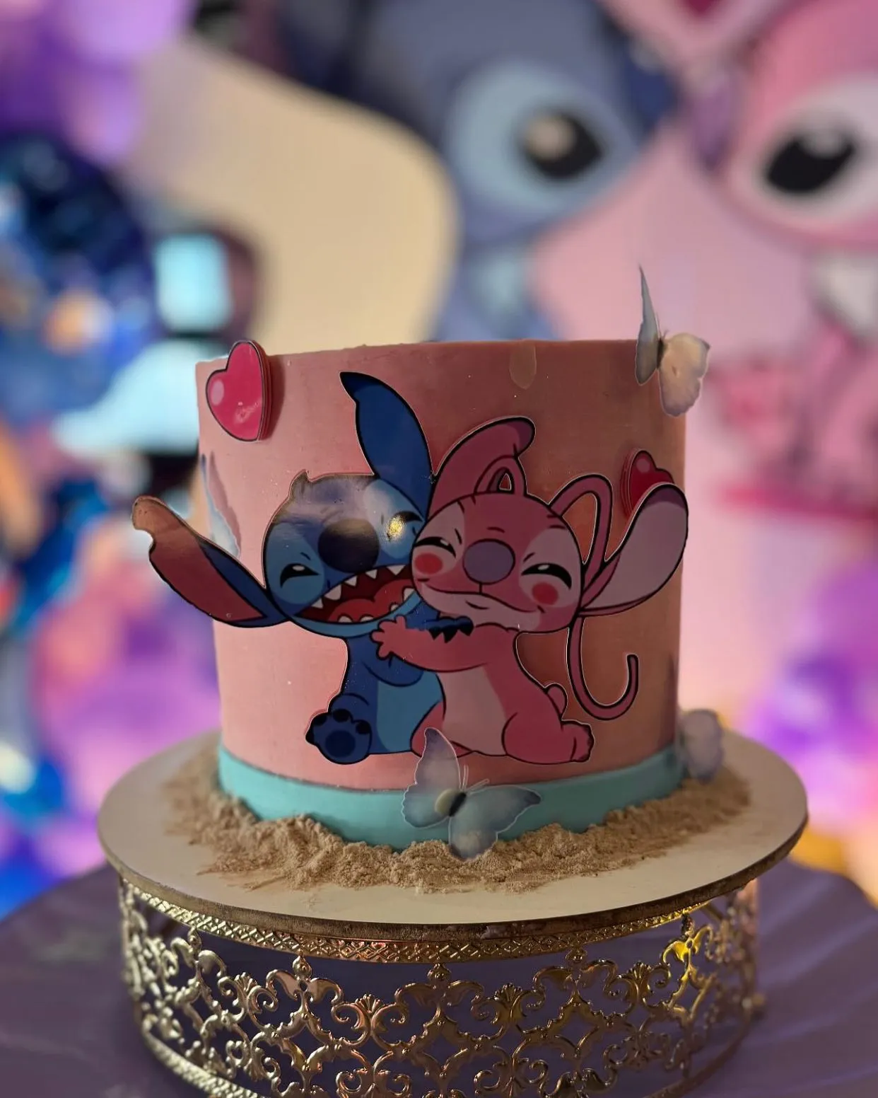 Bolo redondo rosa com uma figura do Stitch abraçando a Angel no topo. A base do doce imita areia e tem uma faixa azul clara.