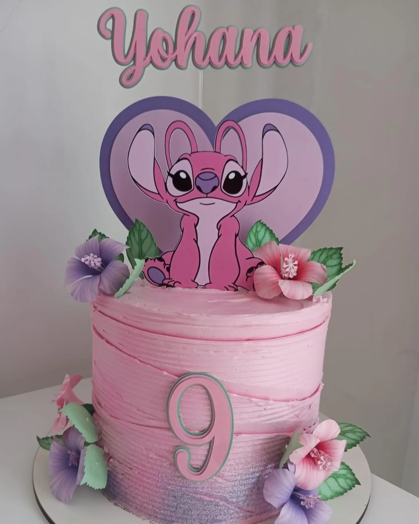 Bolo cilíndrico rosa com glitter lilás na base e o número 9 em destaque. O topo tem um adorno da Angel e flores.
